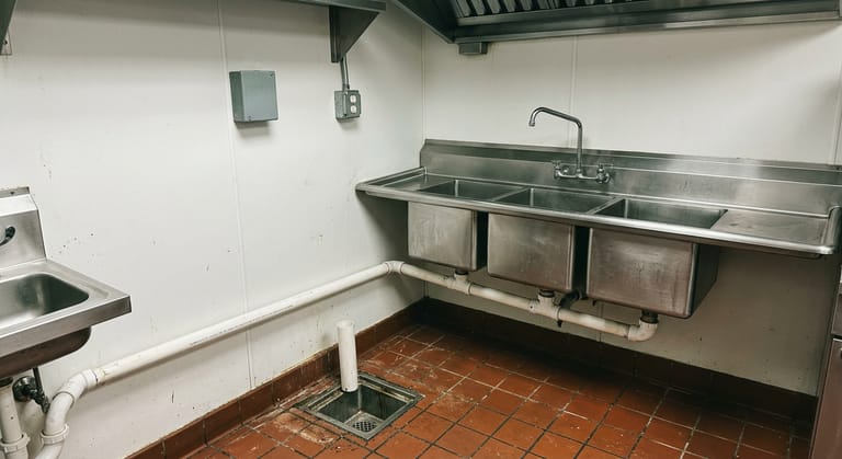 Stainless three-compartment sink in a commercial kitchen with PVC waste piping ending above a floor sink grate.