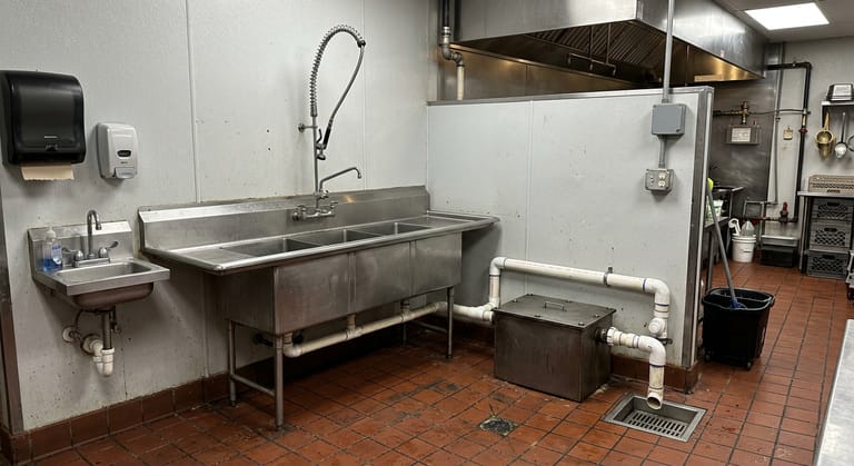 Point of use grease interceptor with a bolted cover on tile beside a commercial three-compartment sink