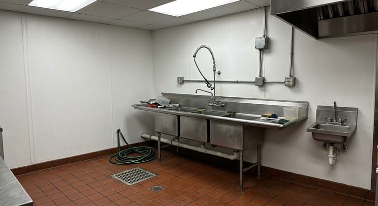Commercial kitchen dish area with a stainless three-compartment sink and a floor sink under an indirect waste pipe.