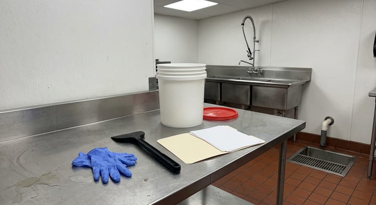 Nitrile gloves, a long-handled scraper, a lidded bucket and a paper file on a commercial kitchen prep counter