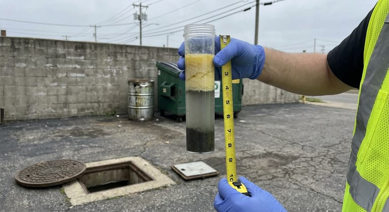 Gloved hand holds a clear sampler tube showing layered grease, water and sludge in a paved service lot.
