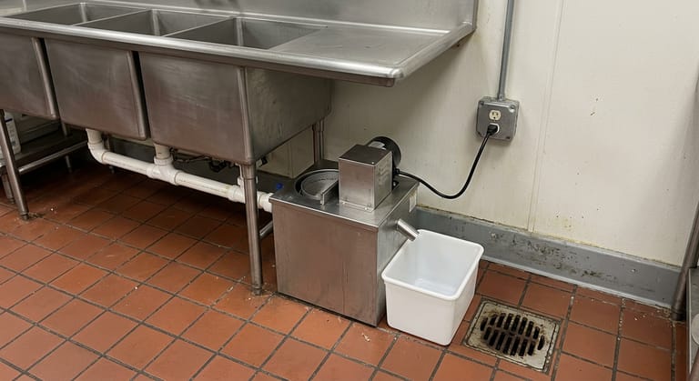 Powered automatic grease removal unit under a commercial sink, empty collection container on the tile beside it.