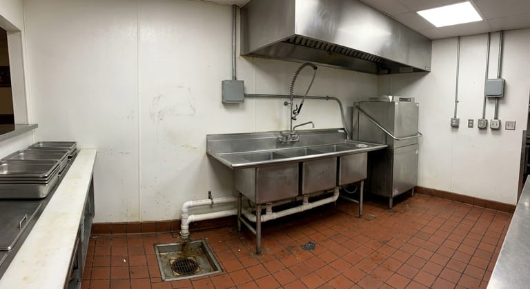 Warewashing corner of a commercial kitchen with a stainless three-compartment sink, dish machine and quarry tile floor.